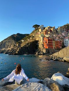 a woman sitting on rocks looking out at the water and buildings in the background,