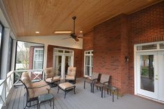 a porch with chairs, tables and ceiling fan on the side of the house that has wood paneling