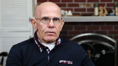 an older man with glasses sitting in front of a fireplace