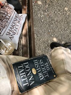 a person holding a book in their lap while sitting on the ground next to a glass of wine