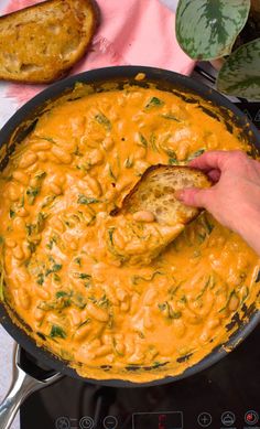 a hand is holding a piece of bread over a skillet filled with cheese and herbs