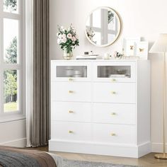 a white dresser sitting next to a bed in a room with large windows and a round mirror on the wall