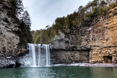 a large waterfall in the middle of a lake surrounded by cliffs and trees on either side