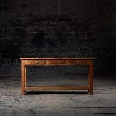 an old wooden table with two drawers on one side and a shelf underneath it, against a dark brick wall