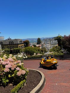 a yellow car driving down a brick road next to flowers and buildings in the background