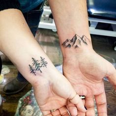 two people holding hands with tattoos on their arms and mountains in the distance behind them