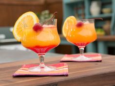 two glasses filled with orange juice and garnished with cherries sitting on top of a wooden table