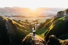 a woman standing on the edge of a cliff looking out over a valley at sunset