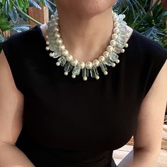 a woman wearing a necklace with pearls and glass beads on her neck, sitting in front of a potted plant