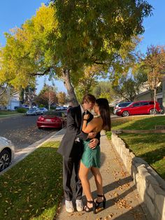 a man and woman kissing in front of a tree on the sidewalk with cars behind them