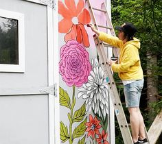 a woman painting flowers on the side of a building with a ladder in front of her