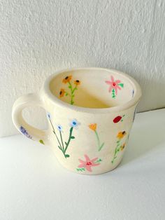 a cup with flowers painted on it sitting on a white counter top next to a wall