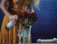 a woman in a yellow dress pouring wine into a vase with red roses on the table