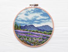 a cross stitch pattern with flowers and mountains in the background on a white wooden surface