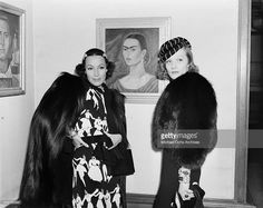 two women standing next to each other in front of a painting on the wall and one wearing a fur coat