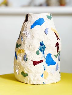 a white vase sitting on top of a yellow table covered in colorful paint splattered paper