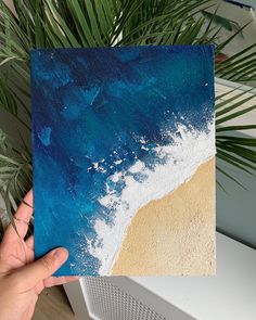 a hand holding up a piece of paper with blue and white paint on it next to a palm tree