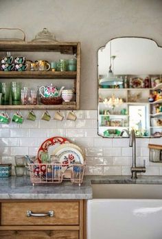 a kitchen sink sitting under a mirror next to a shelf filled with plates and cups