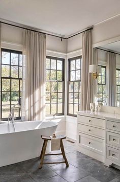 a large white bath tub sitting in a bathroom next to two windows with curtains on them