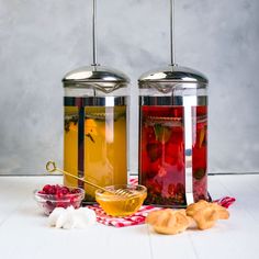 two glass jars filled with liquid next to cookies and cherries on a white table