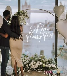 a man and woman standing in front of a window with balloons on it that say, i will you marry me?