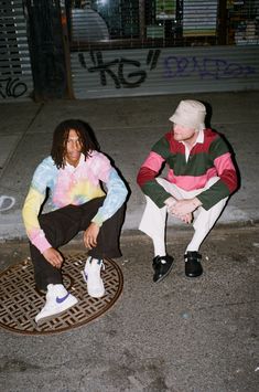 two young men sitting on the ground in front of a manhole cover with graffiti