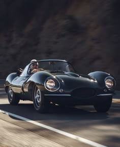 an old black sports car driving down the road in front of a mountain side backdrop