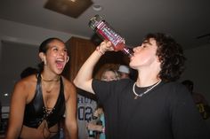 two women drinking from bottles in a room with other people standing around and looking at the camera