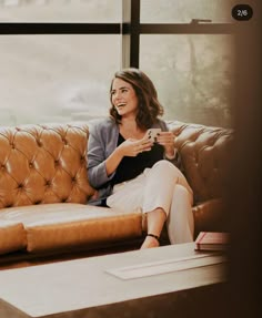 a woman sitting on top of a brown couch next to a window holding a cup