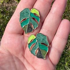 two green leaf shaped brooches sitting on top of a persons hand in the grass