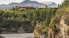 a river flowing through a lush green forest covered hillside next to a tall building with a red roof