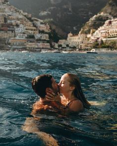 a man and woman kissing in the water next to a town on a cliff side