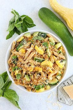 a white bowl filled with pasta and veggies next to a cucumber