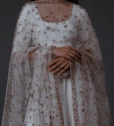 a woman wearing a white gown and cape with gold embroidery on the sleeves, standing in front of a dark background