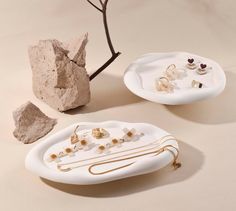 two white plates with jewelry on them next to rocks and a tree branch in the background