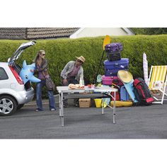 two people standing next to a table with luggage on it and a car in the background