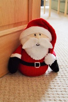 a knitted santa clause sitting on the floor next to a wooden door with his hands in his pockets