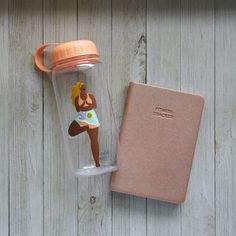 a woman figurine is in a water bottle next to a pink book on a wooden table