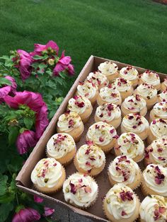 a box full of cupcakes with white frosting and pink flowers in the background