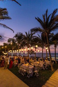 a group of people sitting at tables under palm trees next to the ocean with lanterns hanging from them