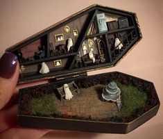 a hand holding an open box with miniature houses in it on top of a table