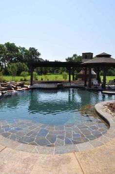 an outdoor swimming pool surrounded by stone pavers and gazebo with seating area in the background