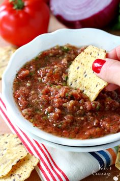 a hand dipping a tortilla chip into a bowl of salsa with tomatoes and onions in the background