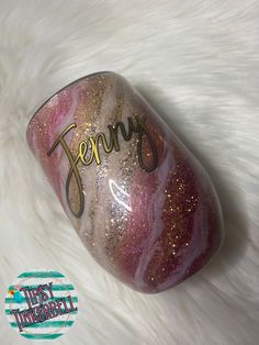 a pink and gold glass with the word jenny on it sitting on a white fur covered surface