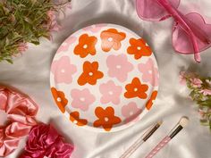 a pink and orange plate with flowers on it next to makeup brushes, flowery hair clips, and other accessories