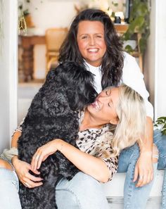 two women sitting on a bench with a black dog in their lap and one woman hugging her