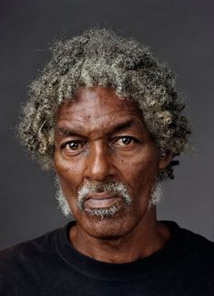 an older man with grey hair and a beard looks at the camera while wearing a black shirt