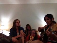 four women are sitting on a bed playing guitar and drinking wine in the same room