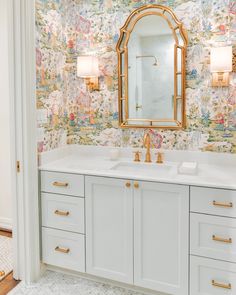 a bathroom with floral wallpaper and gold accents on the mirror above the double sinks