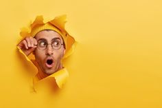 a man wearing glasses and a yellow hat peeks through a hole in the paper